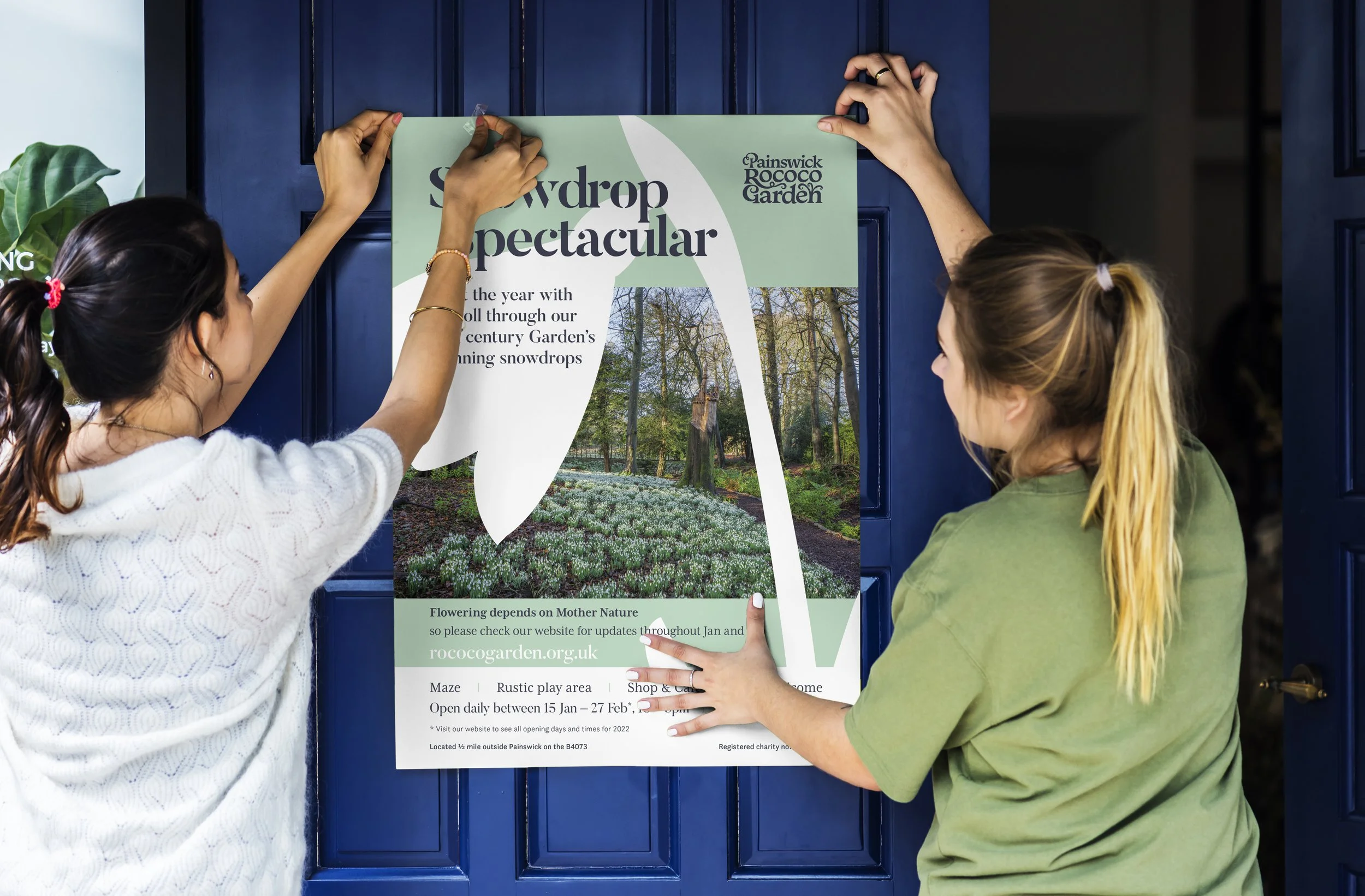 Two women hang up a colorful poster on a blue door. The poster promotes a snowdrop event at Painswick Rococo Garden.
