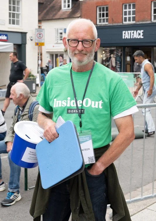 A man with gray hair and a beard wearing glasses and a green t-shirt that says 'Inside Out Dorset' stands on a busy street. He is holding a blue foam pad and a bucket, with lanyard and volunteer badge around his neck. Several people in the background are walking and sitting.