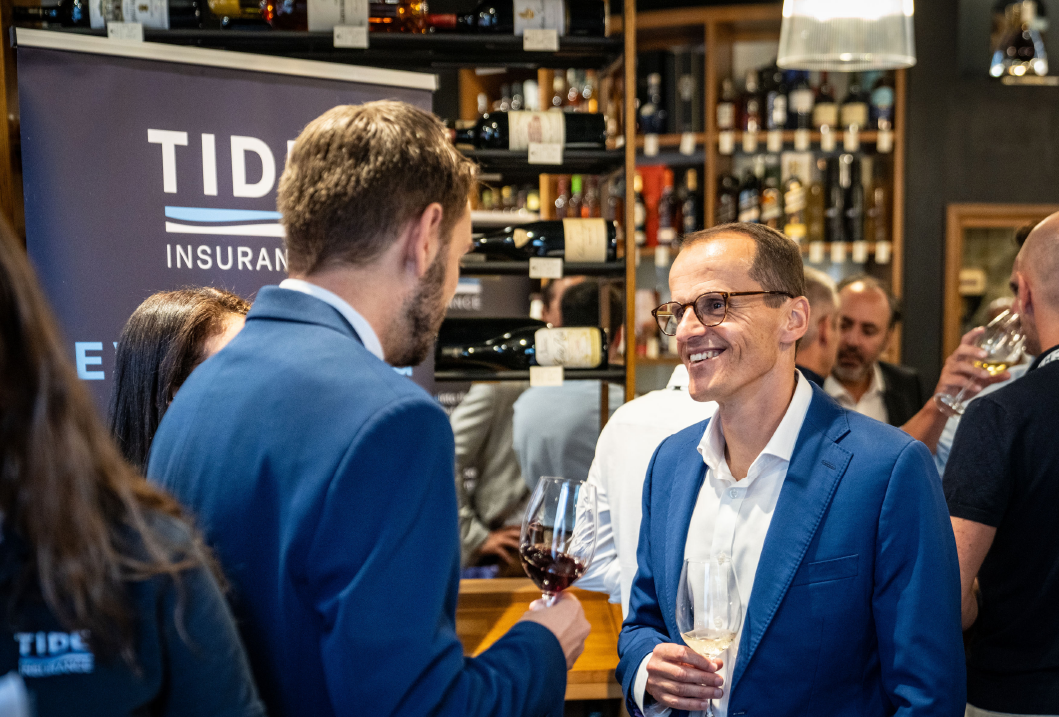 People at a social event in a wine bar, wearing formal attire, holding wine glasses, with shelves of wine bottles in the background.