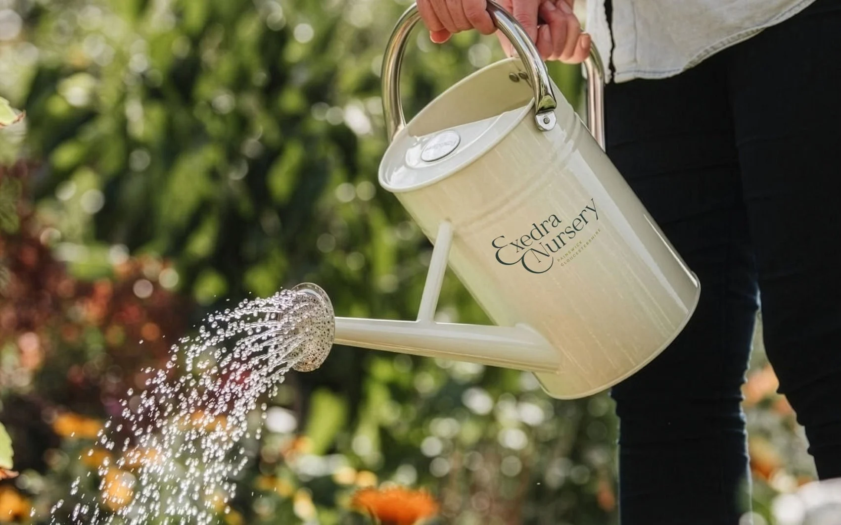 Person watering plants with a cream watering can labeled 'Exedra Nursery' outdoors in a garden.