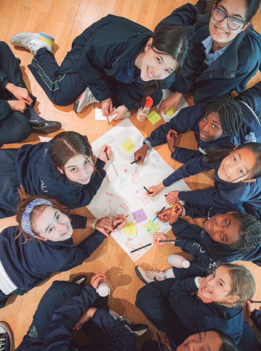 Group of diverse schoolchildren sitting on a wooden floor, working together on a large colorful poster with sticky notes and markers, smiling and collaborating.