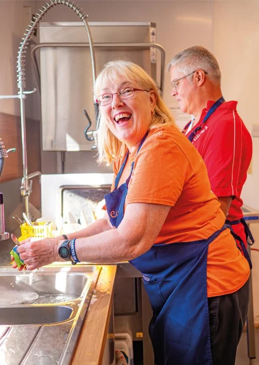 A woman with glasses laughing while washing dishes at a kitchen sink, with an older man in red in the background in a commercial kitchen setting.