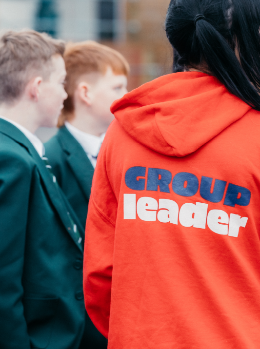 A person wearing an orange hoodie with the words 'GROUP leader' on the back, standing in front of two children dressed in school uniforms.