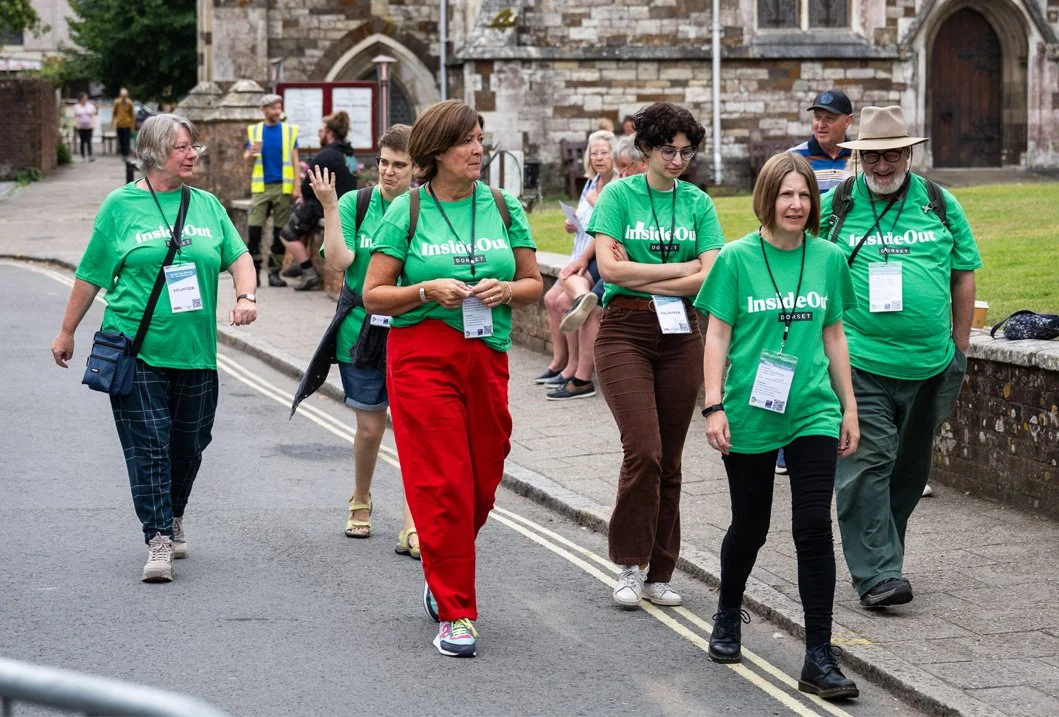 Group of people walking and talking outdoors, some wearing green 'Inside Out' shirts and badges, near historic stone buildings.