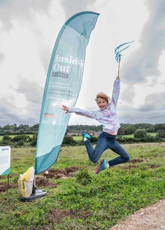 A young girl jumping in the air next to a blue festival banner outdoors on a cloudy day.
