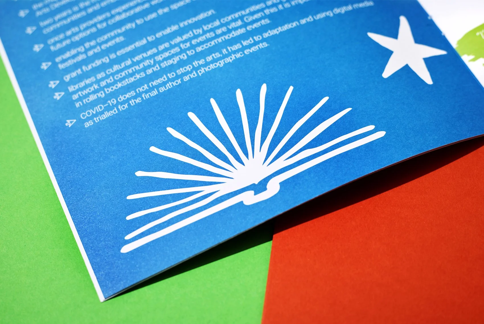 Close-up of a blue printed sheet of paper with white graphic of an open book and stars, placed on green and red papers, with some text visible.