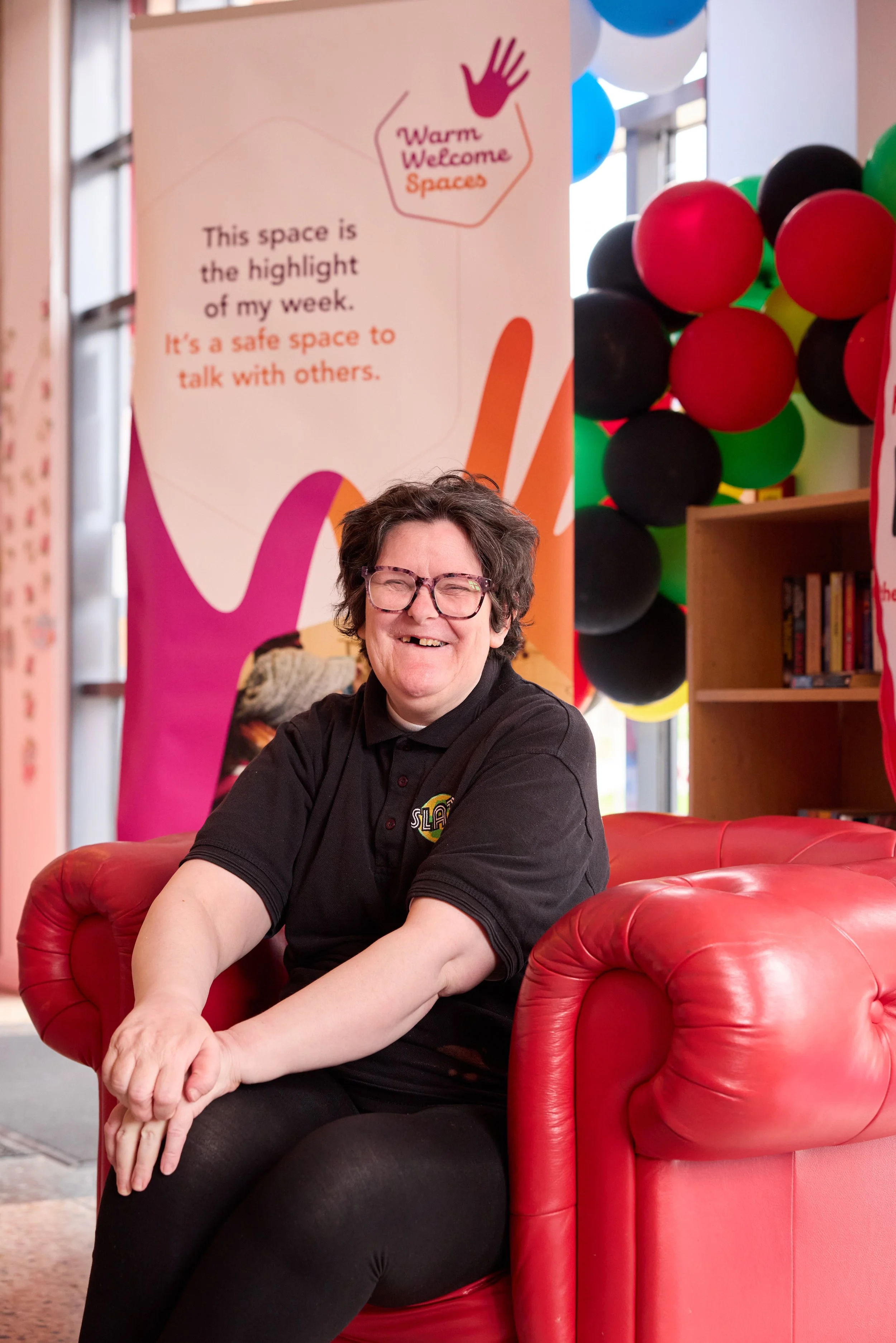 A smiling woman with glasses sitting on a red leather couch, with a colorful balloon display and a sign that reads "Warm Welcome Spaces" behind her.