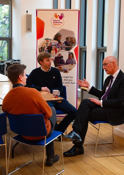 Three men having a conversation in a room with large windows and a colorful banner that reads 'Warm Welcome Spaces' in the background.