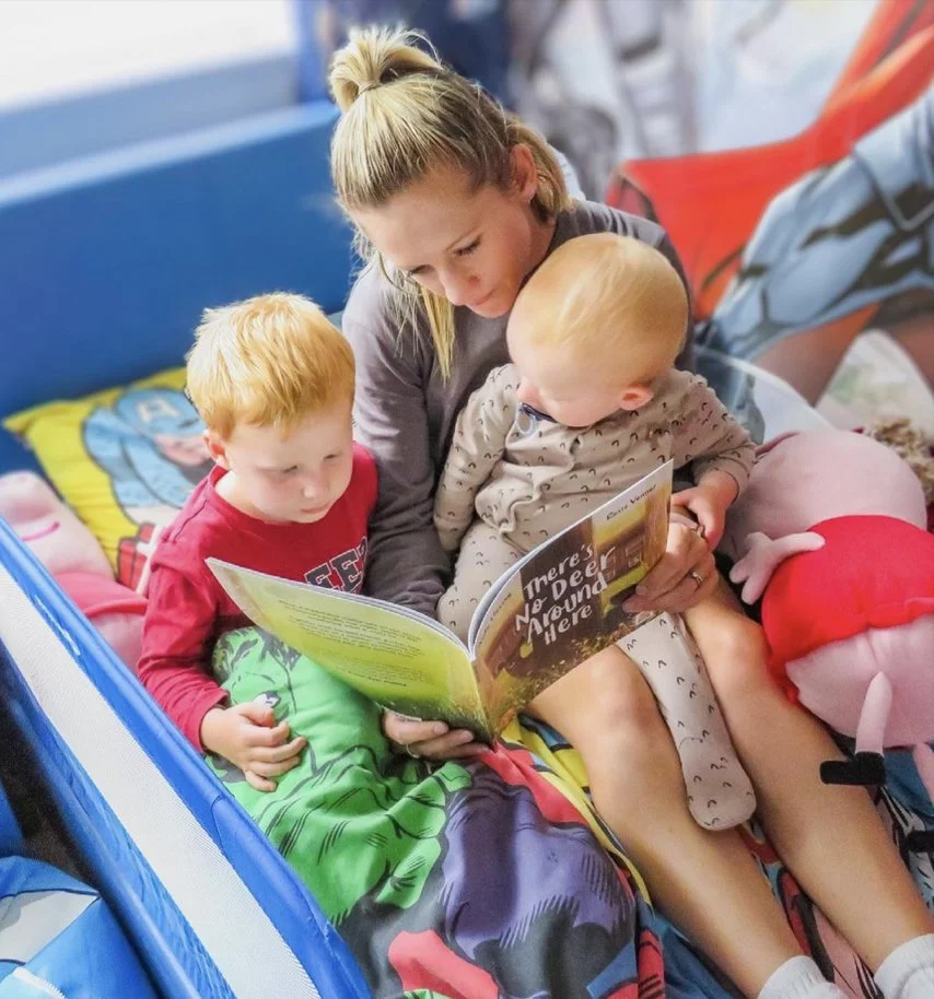 A woman reading a children's book to two young kids, one with red hair and another with blonde hair, seated on a bed with colorful bedding and pillows in a lively room.
