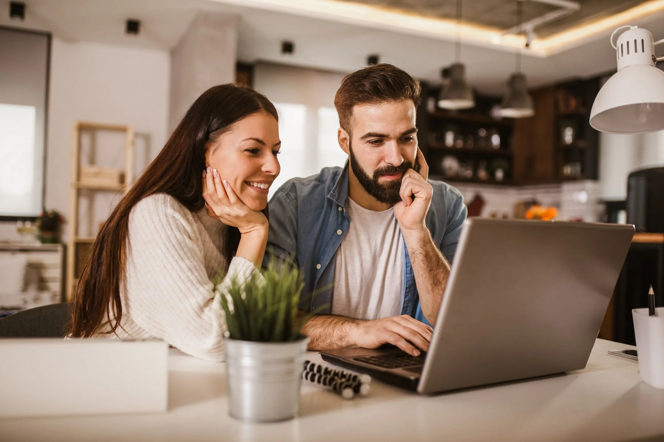 A young woman and a young man sit together at a table, looking at a laptop screen, with the woman smiling and resting her chin on her hand, and the man concentrating with his hand on his face. The background shows a cozy, modern kitchen.