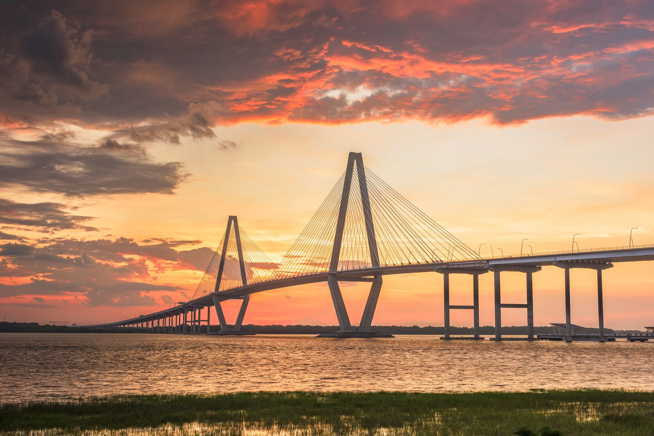 A modern cable-stayed bridge spanning over a body of water at sunset, with clouds in the sky.