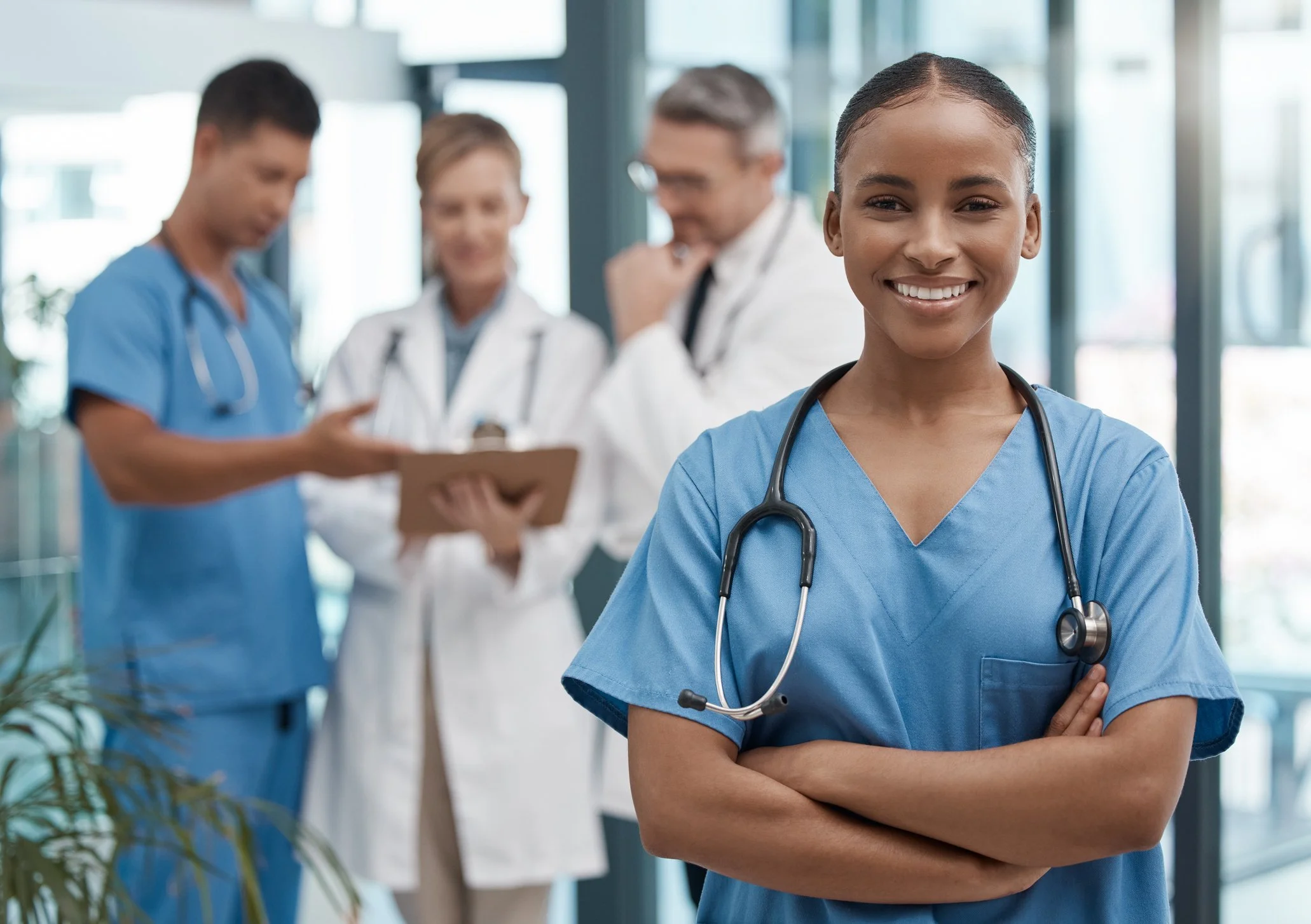 Smiling female nurse in blue scrubs with a stethoscope around her neck, standing confidently with arms crossed in a hospital or medical facility, with four other healthcare professionals in the background.