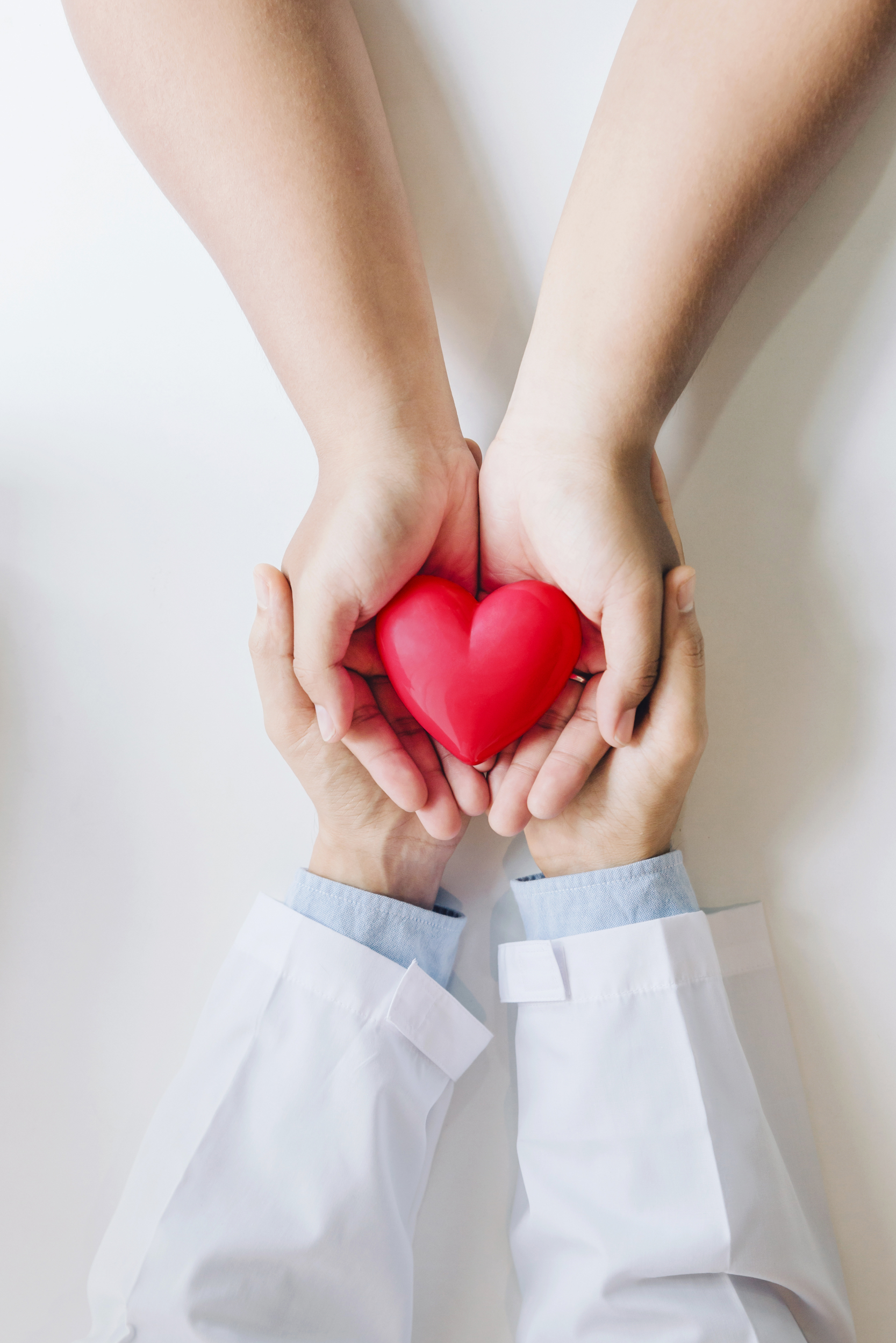 Two hands holding a red heart-shaped object, with two other hands underneath supporting it; one person's arms are in a white coat, suggesting a healthcare professional.