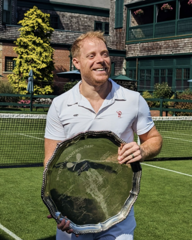 Camden Riviere, world real tennis champion, smiling and holding a silver tennis trophy on a tennis court. He is wearing a white polo shirt and blue shorts, and the background features a net, a green building with balconies, and trees.