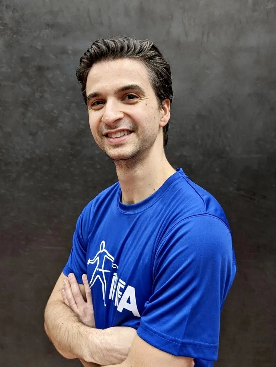 Dan Tristao, Rugby Fives champion. A young man with dark hair, smiling, wearing a blue T-shirt with a white logo, standing with arms crossed against a dark background.