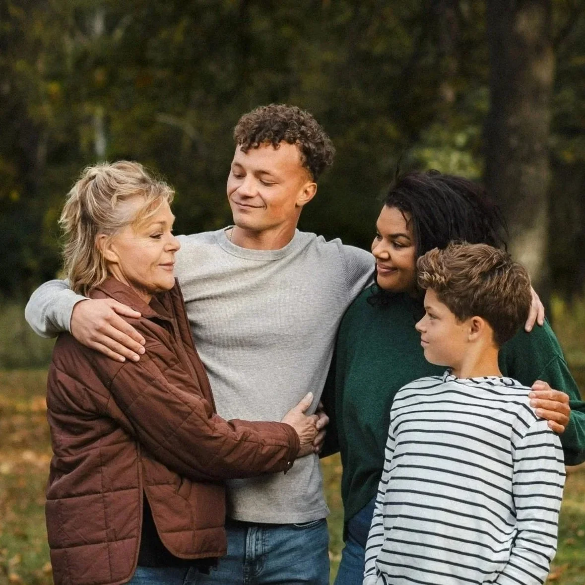 Four people, an elderly woman, two young women, and a young boy, embrace outdoors in a park-like setting, sharing a tender moment.