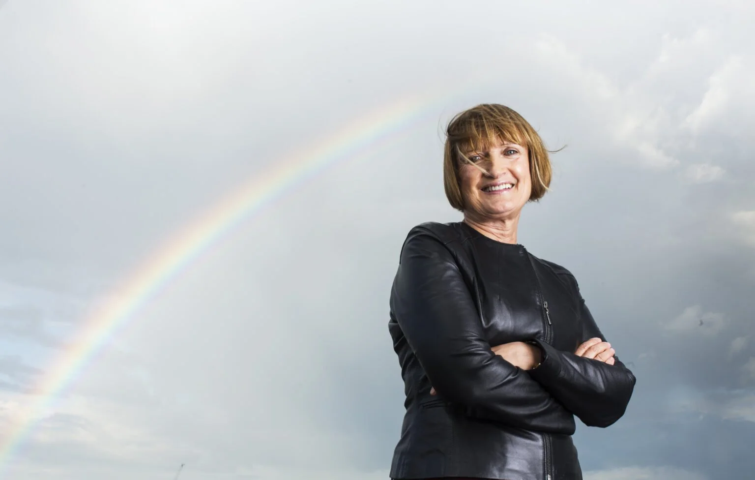 A smiling aTessa Jowell wearing a leather jacket standing outdoors with crossed arms, under a cloudy sky with a rainbow in the background.