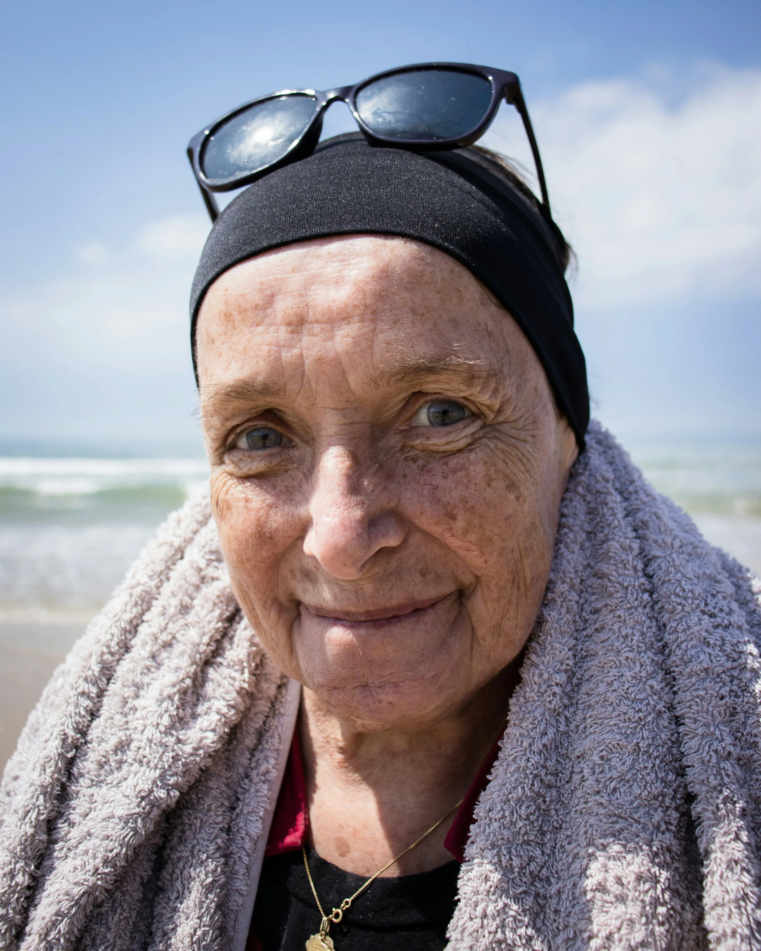 Close-up of an elderly woman with grey hair, blue eyes, wearing sunglasses on her forehead, a black headband, a dark shirt, and a towel around her shoulders, at the beach with the ocean in the background.