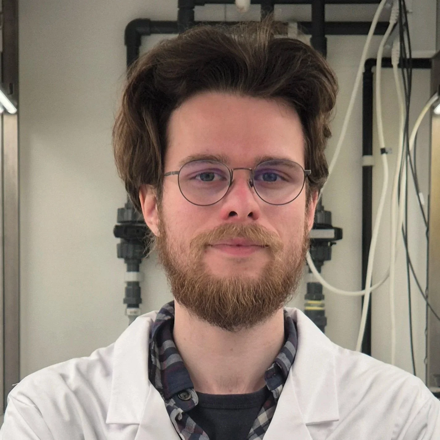 A young man with glasses and a beard, wearing a white lab coat, takes a selfie in a laboratory environment.
