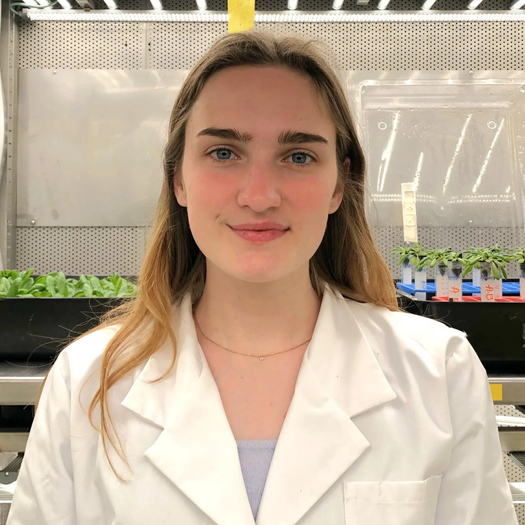 A young woman with light skin, long light brown hair, and blue eyes, wearing a white coat and a delicate gold necklace, standing in a laboratory or greenhouse environment with trays of plant seedlings in the background.