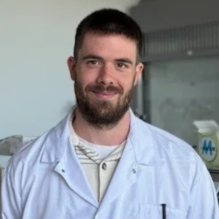 A young man with a beard and short dark hair, wearing a white medical coat, smiling in a clinical setting.