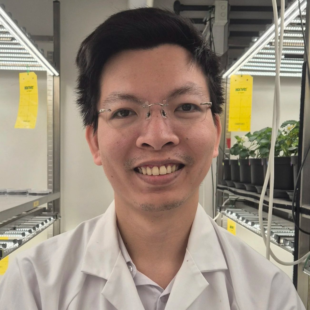 A smiling man with glasses and a white lab coat standing in a laboratory with plants and equipment in the background.