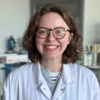 A smiling woman with curly brown hair and glasses, wearing a white lab coat, standing in a medical or laboratory environment.