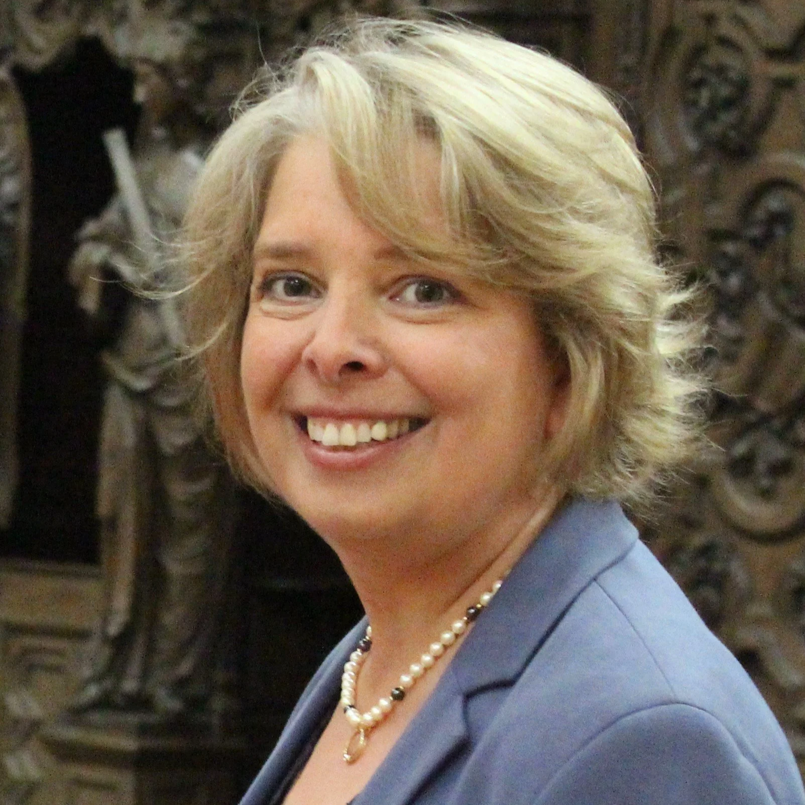 Close-up of a smiling woman with short blonde hair, wearing a pearl necklace and a blue blazer, with a dark ornate background.