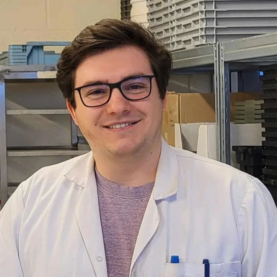 Young man with dark hair and glasses smiling, wearing a lab coat, in a laboratory or storage room with shelves and boxes in the background.