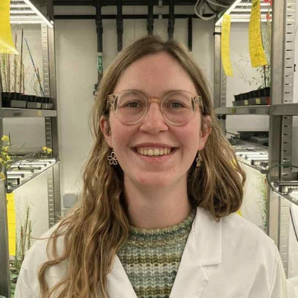 A young woman with long wavy hair, glasses, and earrings, smiling in a laboratory or greenhouse setting with plants and shelves in the background.