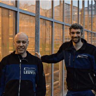 Two men standing in front of a glass building, smiling, wearing dark jackets with blue accents.