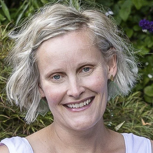 A woman with short blond hair smiling outdoors with green plants and purple flowers in the background.