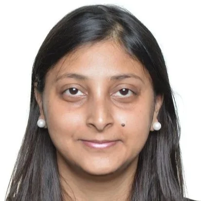 Close-up of a woman with long black hair, wearing pearl earrings, and a small mole on her right cheek, looking at the camera against a white background.