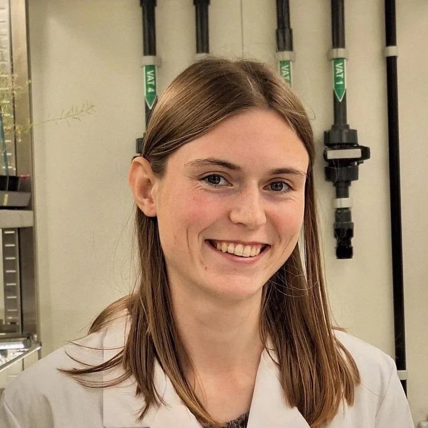 Young woman with long brown hair smiling in a laboratory or medical setting.