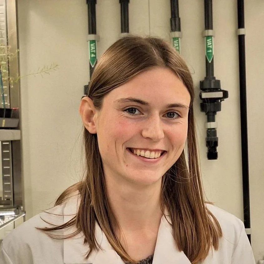Young woman with shoulder-length brown hair smiling in front of laboratory equipment, including several vertical pipes or tubing.