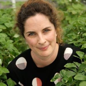 A woman with curly brown hair outdoors surrounded by green plants, wearing a black top with white and peach-colored patterns, smiling at the camera.