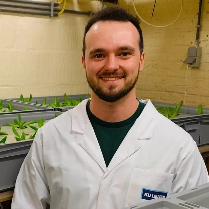 A man in a white lab coat stands in a room with green plants growing in gray containers.