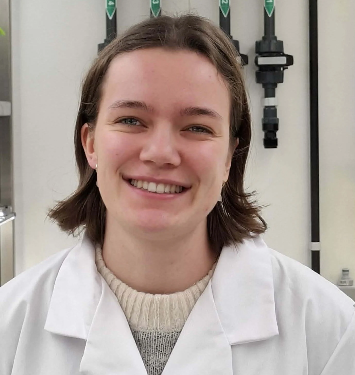 A young woman with short brown hair and light skin, smiling, wearing a white lab coat and a beige sweater, in a laboratory setting.