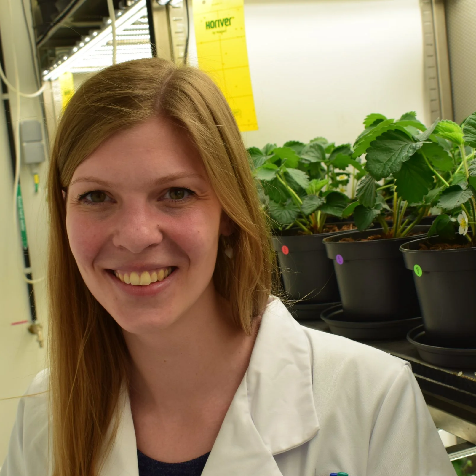 A smiling woman with long red hair in a lab coat standing near potted plants labeled with colored stickers inside a laboratory.
