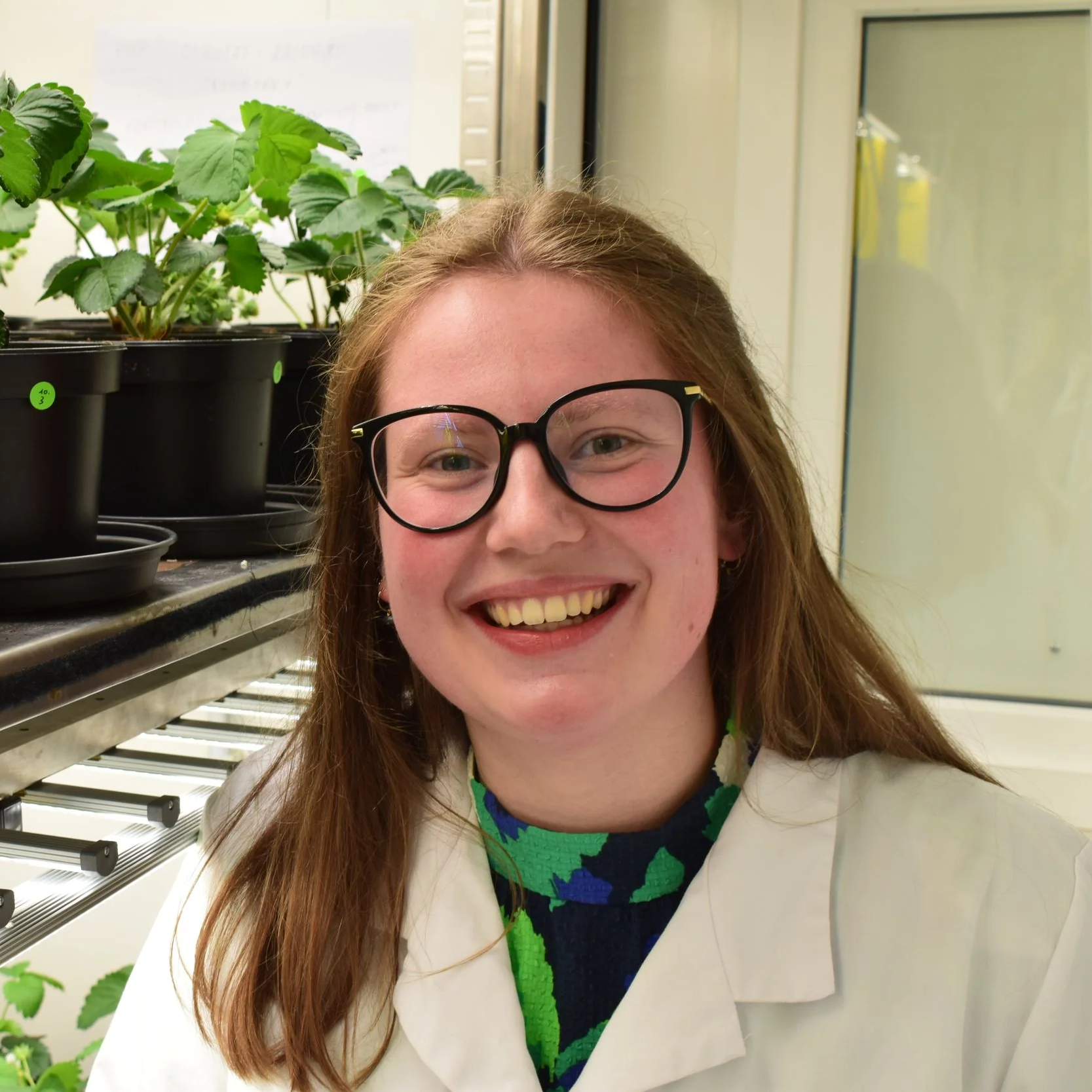 A young woman with red hair, glasses, and a bright smile wearing a white lab coat and colorful shirt, standing in a laboratory with potted plants and scientific equipment.