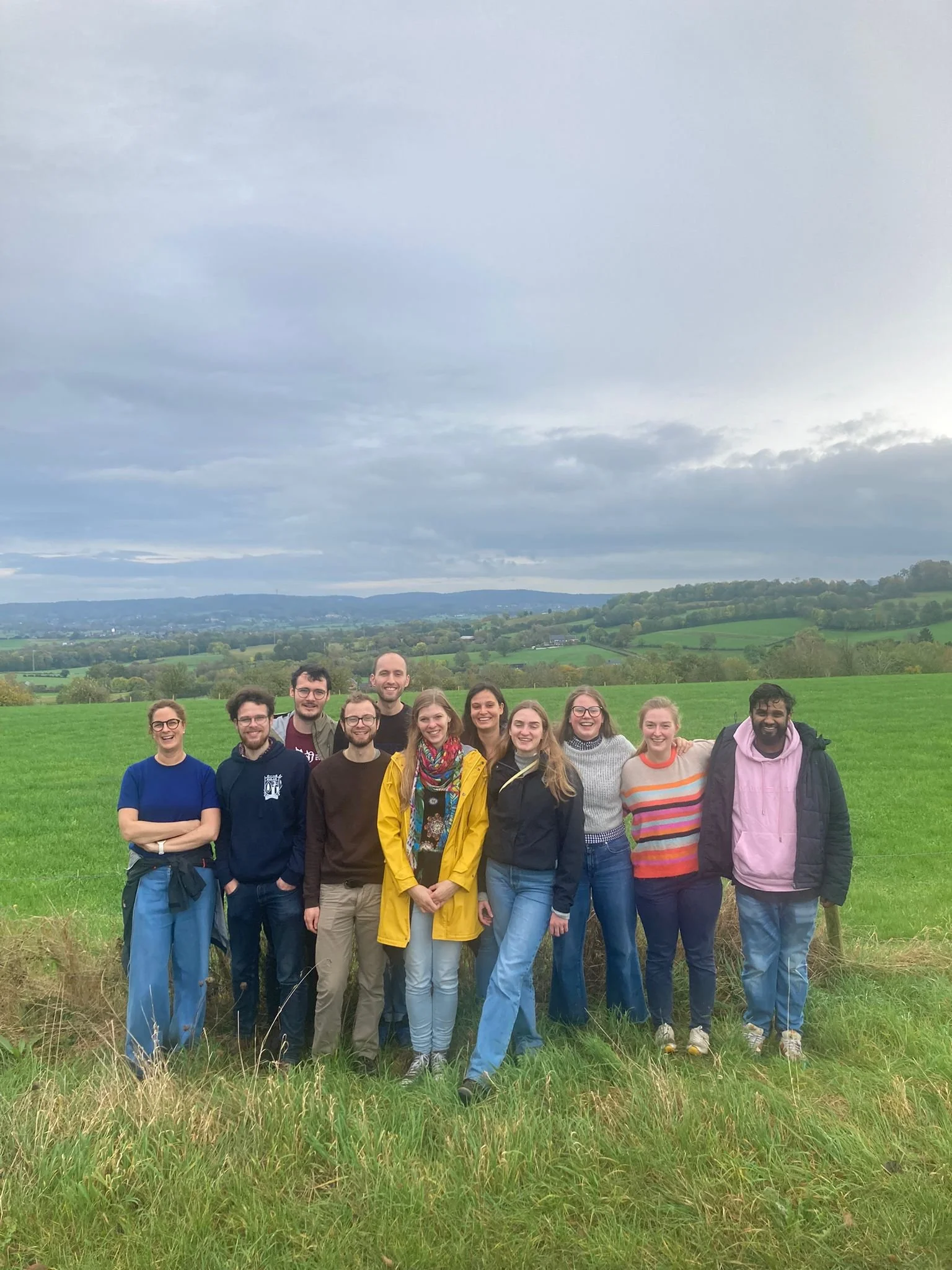 Group of eleven people smiling and standing together outdoors on a grassy field with a scenic landscape of rolling hills and cloudy sky in the background.