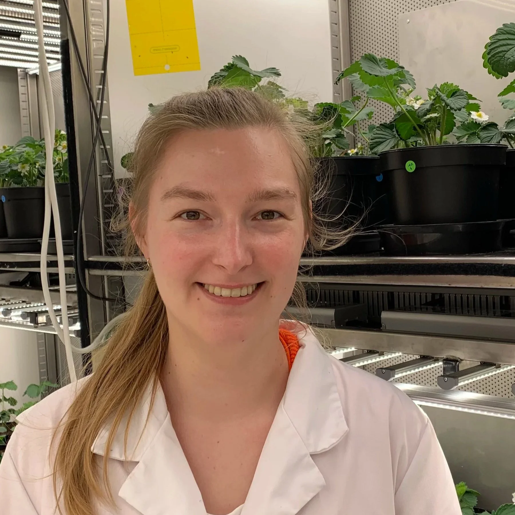 A woman with long blonde hair smiling in a laboratory or greenhouse with potted plants in the background.