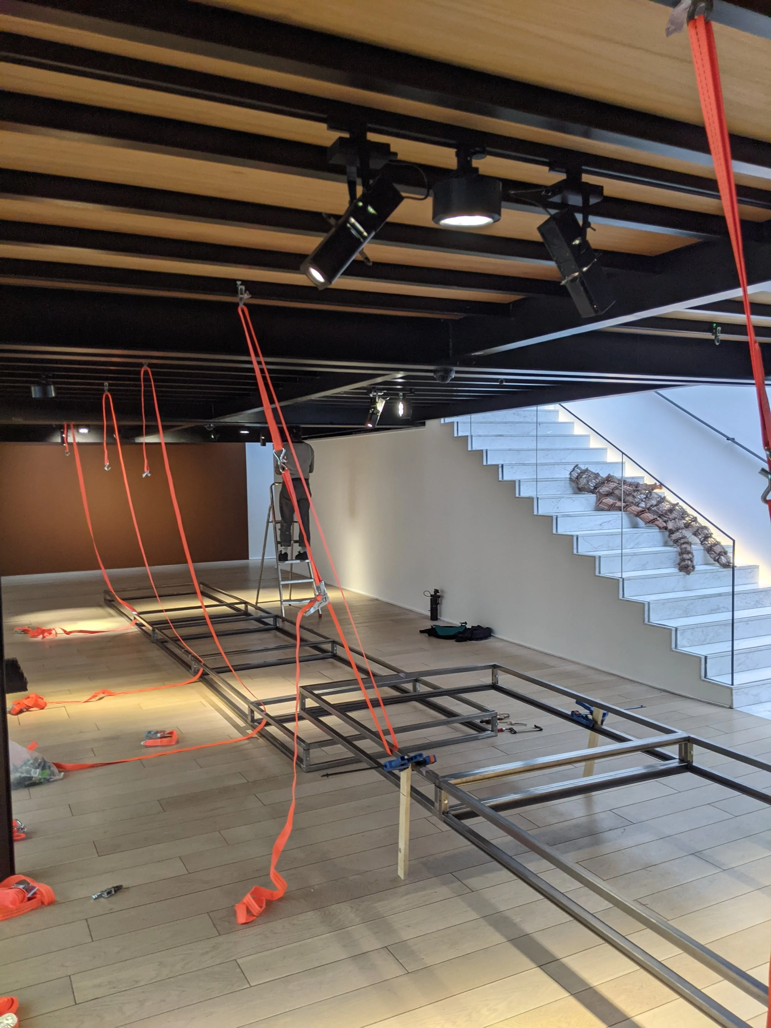 Une œuvre en cours de montage de Kenny Dunkan, avec une structure métallique sur le sol, des sangle oranges suspendues, un escabeau, et une escale avec des chaussures ou vêtements dessus, près de marches en marbre menant à un étage supérieur. 