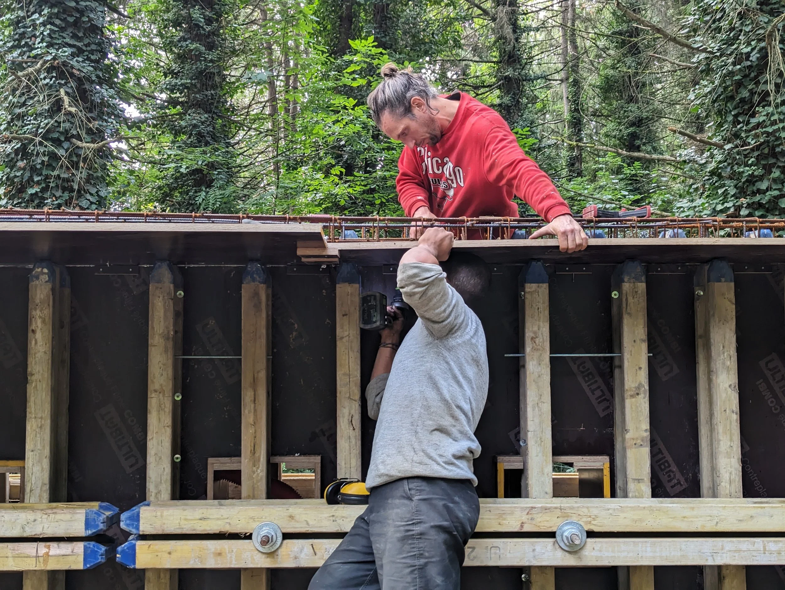 Deux hommes travaillent sur une structure en bois dans une forêt pour l'installation "Vestiges" d'Hicham Berrada. L'un est allongé en dessous et l'autre se tient au sommet de la structure.