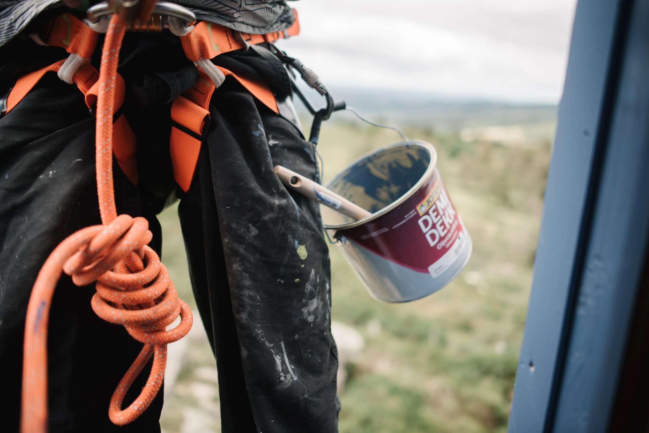 Une personne en équipement d'escalade, portant un harnais orange, avec un seau de peinture suspendu à leur côté pour la rénovation du "Phare" de Gloria Friedman pour le Parc Naturel Régional des Monts d'Ardèche.