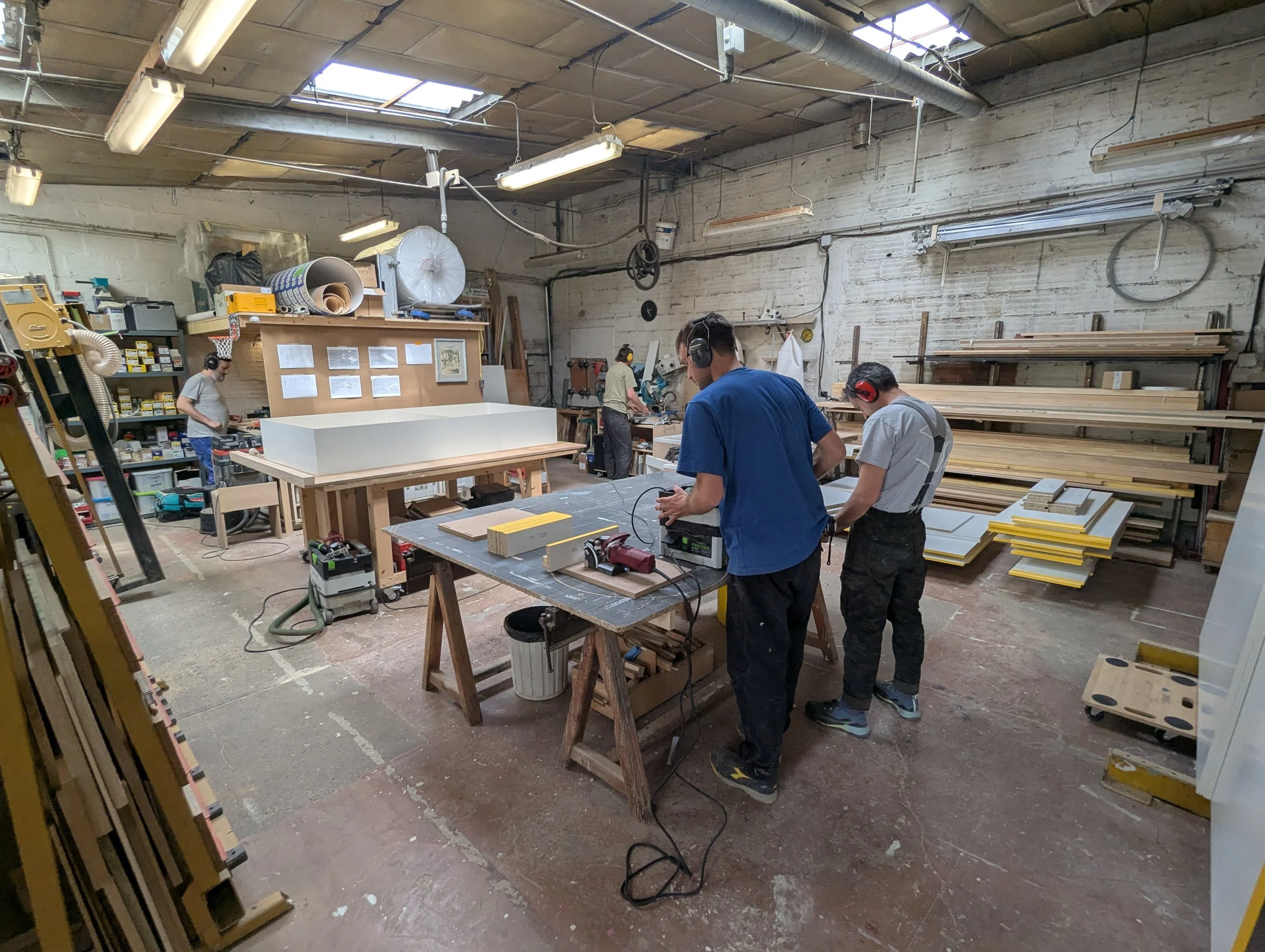 Des artisans travaillent dans un atelier de menuiserie, utilisant des outils électriques, avec des étagères de planches en bois, et portant des protections auditives. Atelier Socle situé à Vitry-sur-Seine aux portes de Paris.