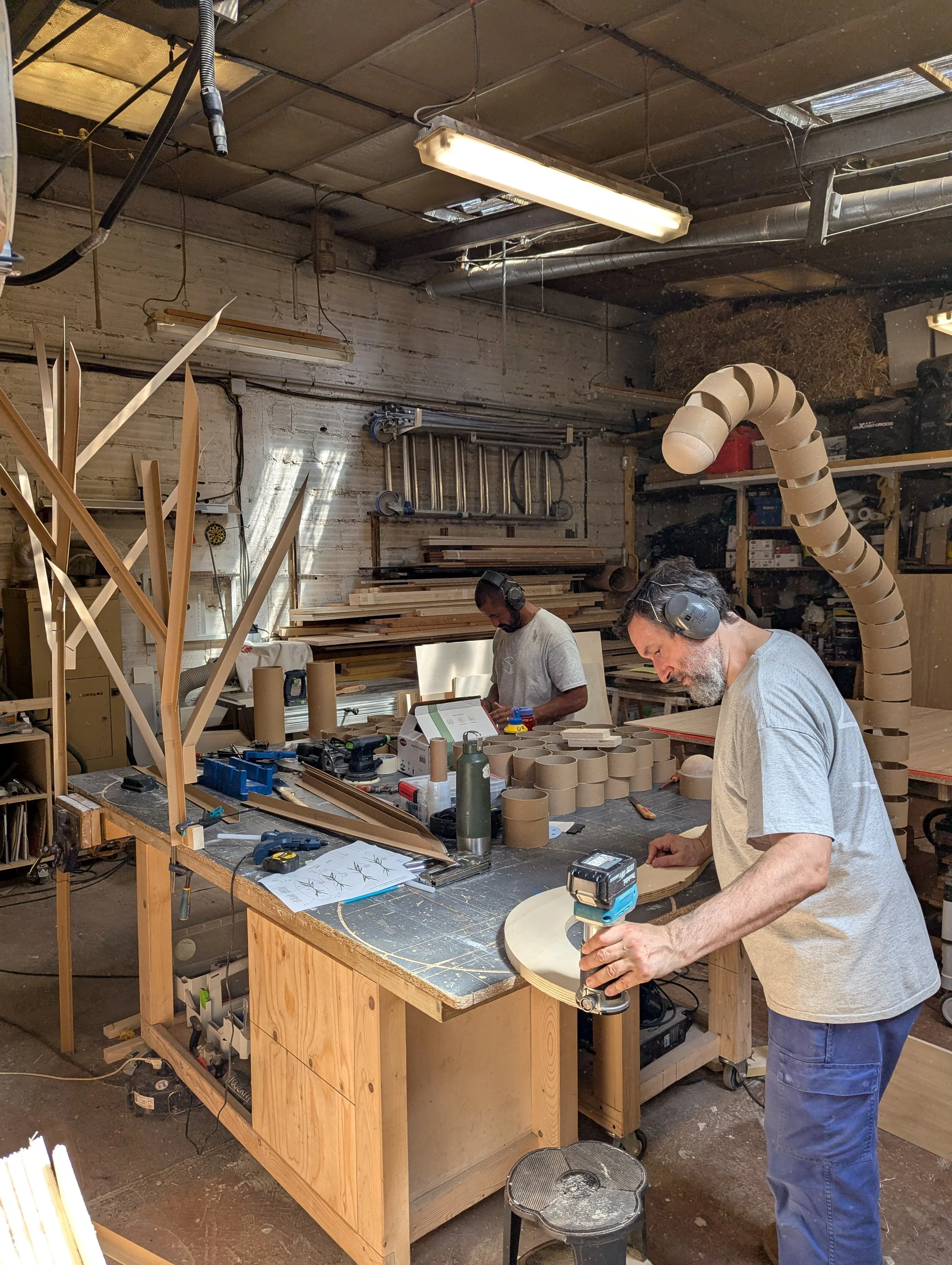 Deux hommes travaillent dans un atelier de menuiserie, utilisant des outils pour fabriquer des objets en bois, entourés de planches, de rouleaux de papier, et divers matériaux de travail. Ils sont à Vitry-sur-Seine en Ile-de-France.