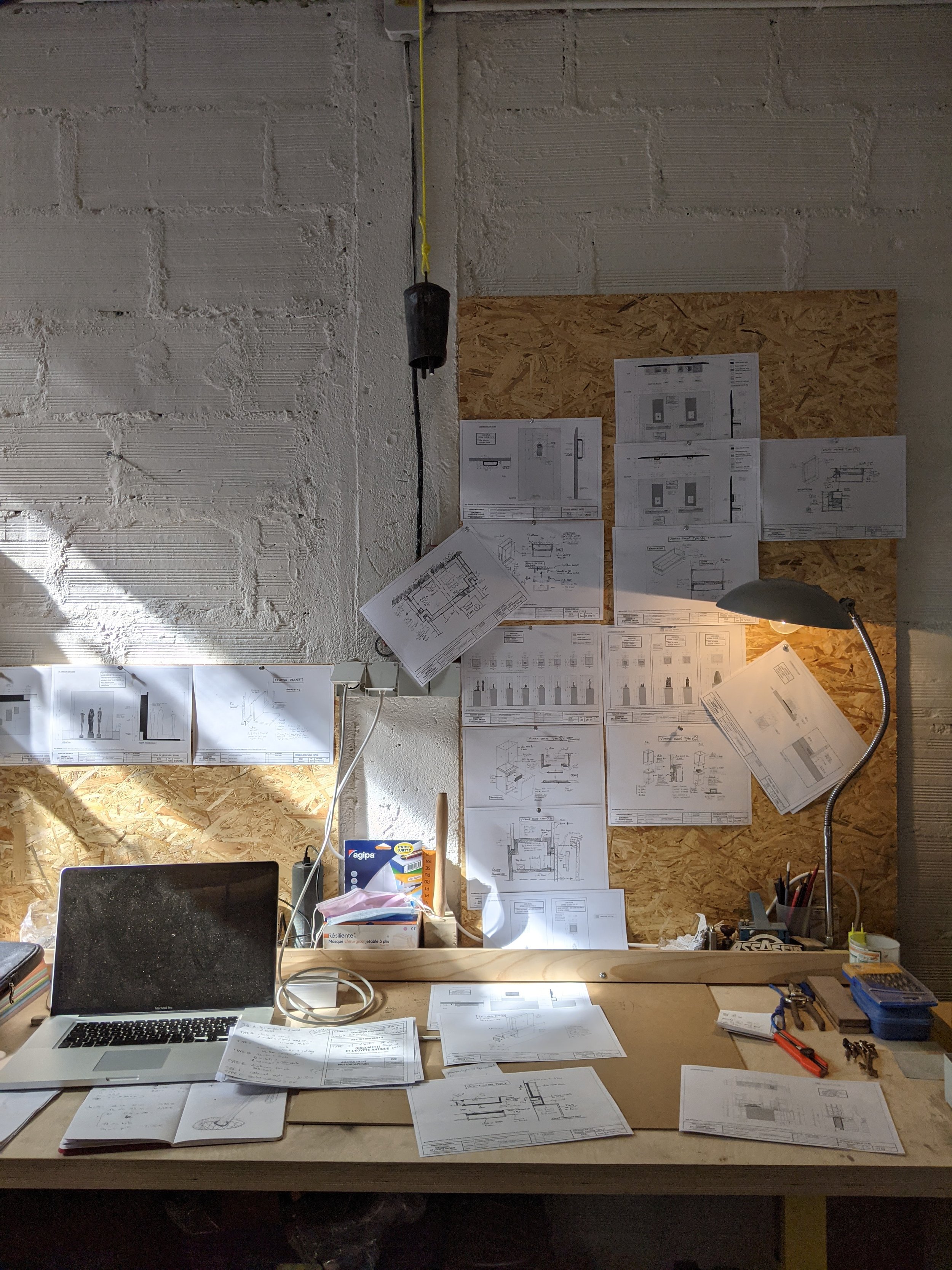 Un plan de construction sur un mur d'atelier, avec une table de travail, un ordinateur portable, des lampes et des documents liés au projet. Cette image illustre la phase conception.
