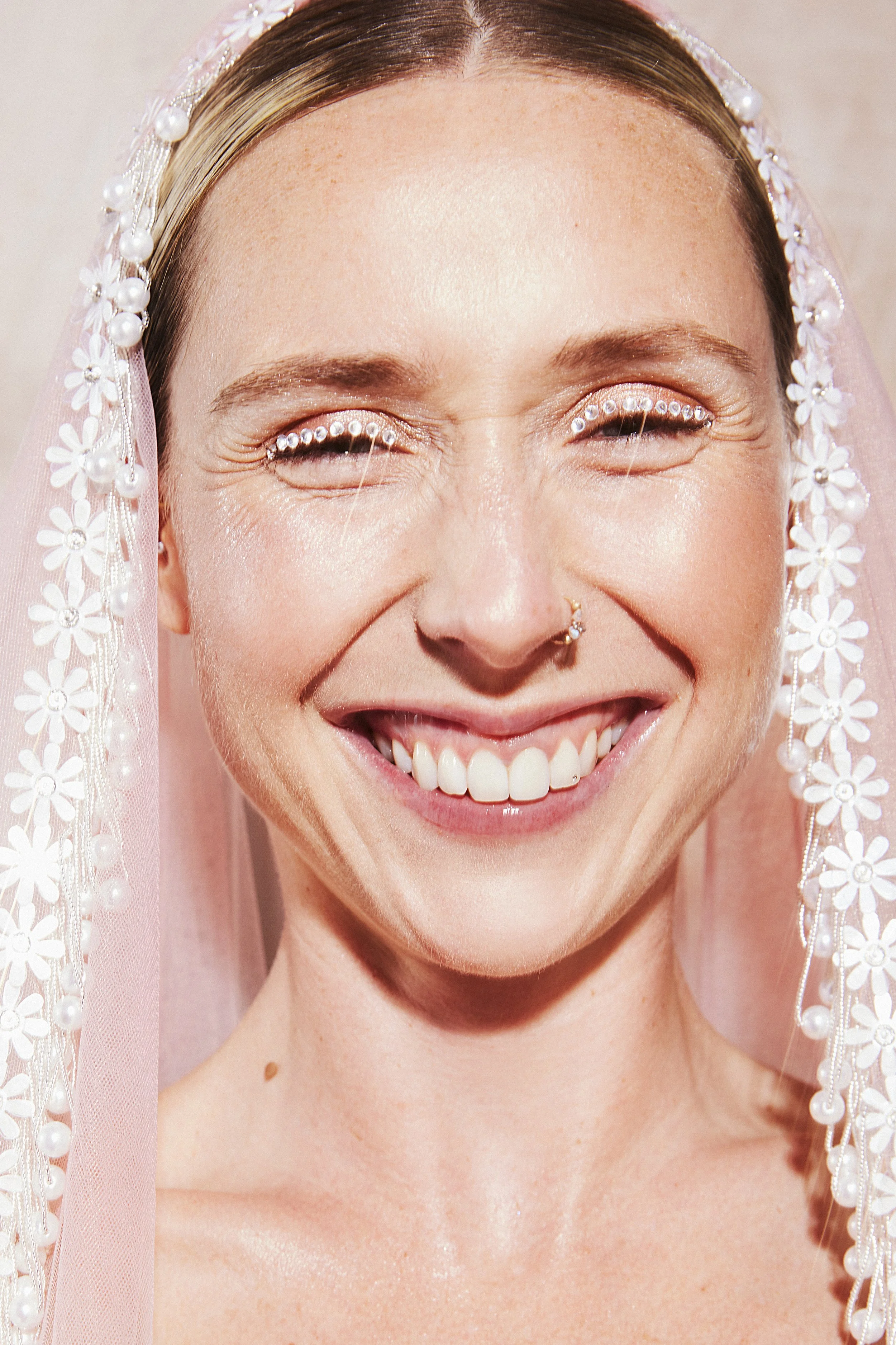 Close-up of a smiling woman wearing a white floral veil with pearls, with face makeup and a nose piercing.