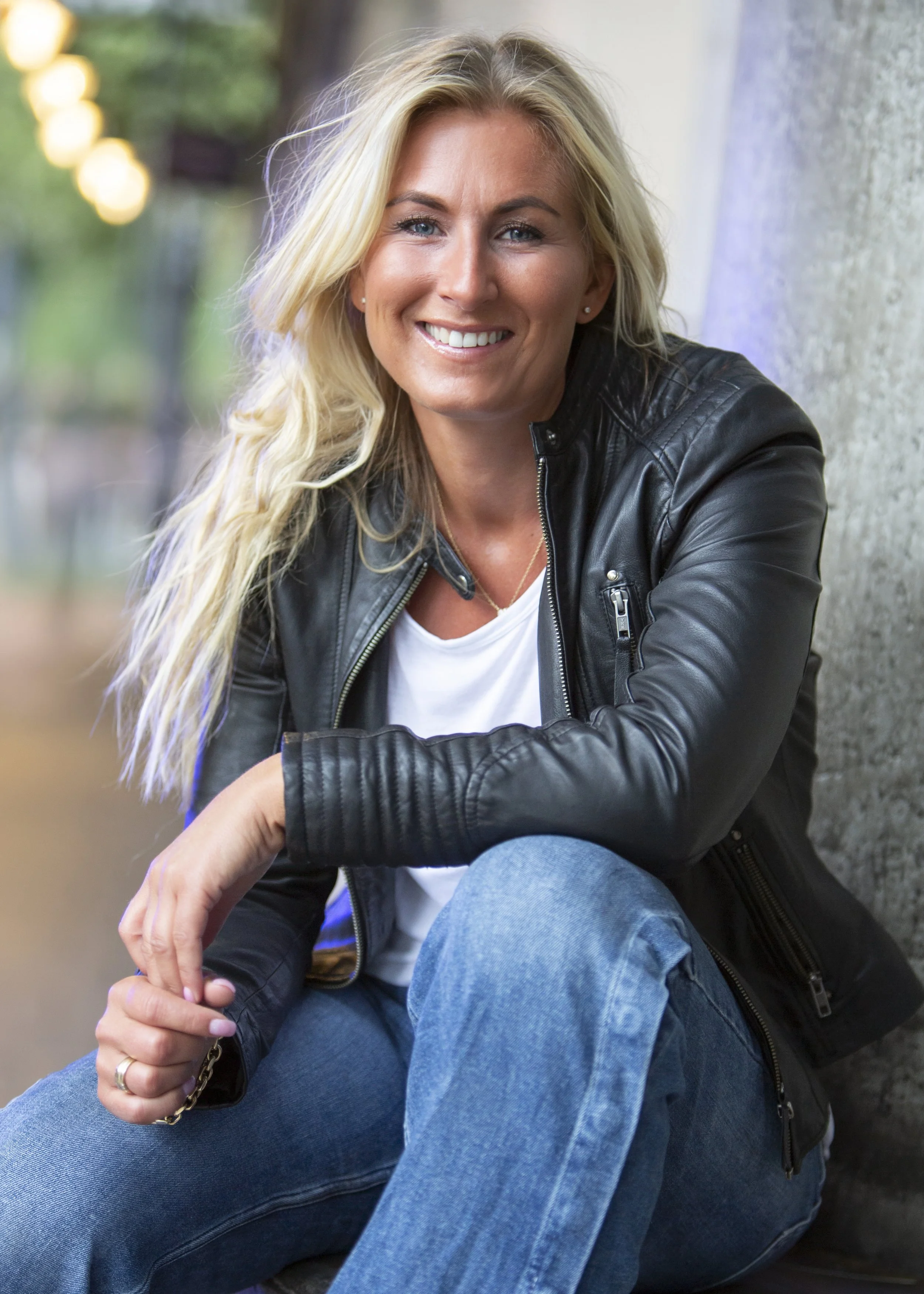A woman with long blonde hair smiling, wearing a black leather jacket and blue jeans, sitting outdoors near a concrete wall.